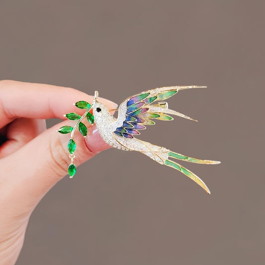 Hand holding a decorative hummingbird brooch with gemstones against a neutral background