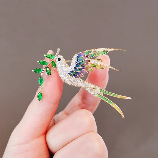 Gold hummingbird brooch with gemstones held by a hand against a neutral background