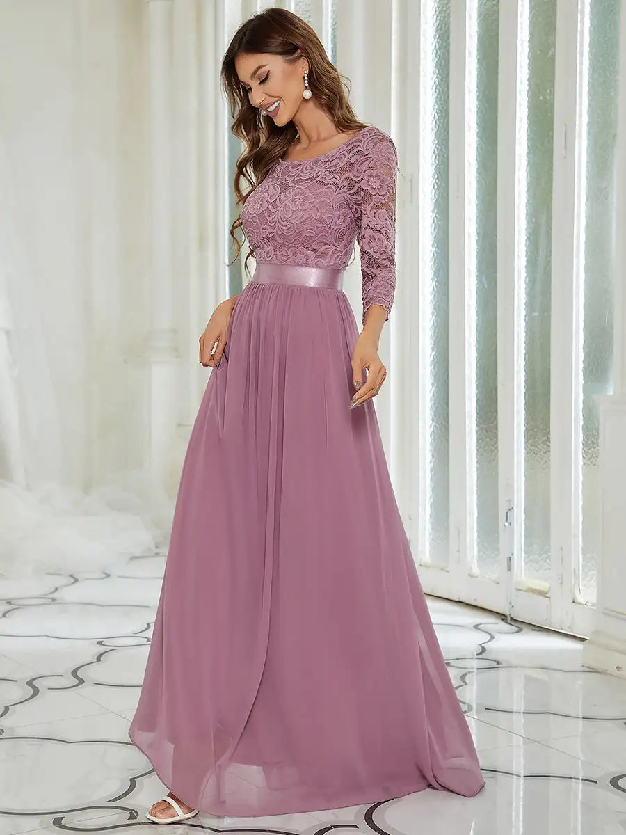 Woman wearing a long, elegant purple dress with lace top and flowing skirt in a bright room.